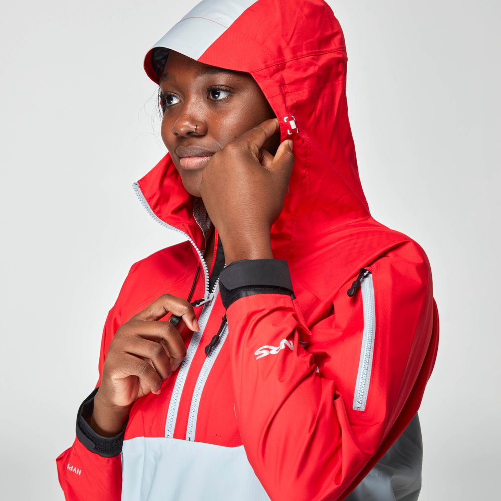 A woman wearing the NRS High Tide Jacket in red and gray zips up the collar and adjusts the hood, facing slightly left against a plain background.