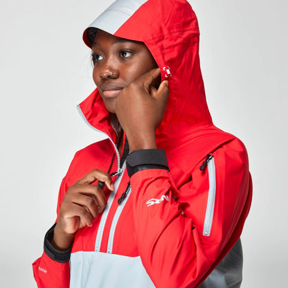 A woman wearing the NRS High Tide Jacket in red and gray zips up the collar and adjusts the hood, facing slightly left against a plain background.