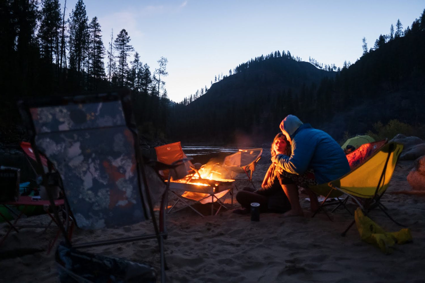 Two people enjoy a campfire at dusk on a sandy riverbank, with empty chairs nearby and trees silhouetted in the background, all using 4CRS Rental Camp Gear.