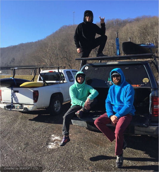 Three people in hooded jackets, including the Immersion Research Hot Lap Hoodie - Men's and a lightweight fleece, sit and stand on a pickup truck by surfboards. It's a sunny day in the mountains—ideal for relaxing outdoors.