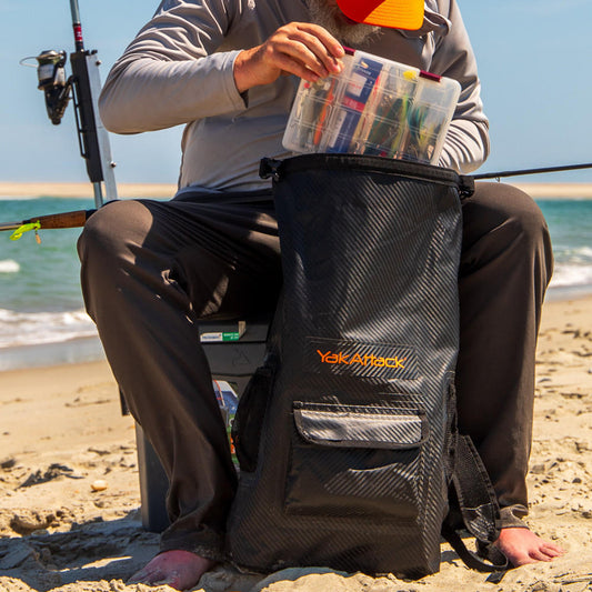 A person sits on the beach by a fishing pole, packing a tackle box into the YakAttack 20L Roll-Top Drybag Backpack—ideal for outdoor adventures near water.