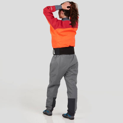A person wears a vibrant red and orange NRS REV (GORE-TEX Pro) Drytop and gray pants, adjusting their hair against a plain light background.