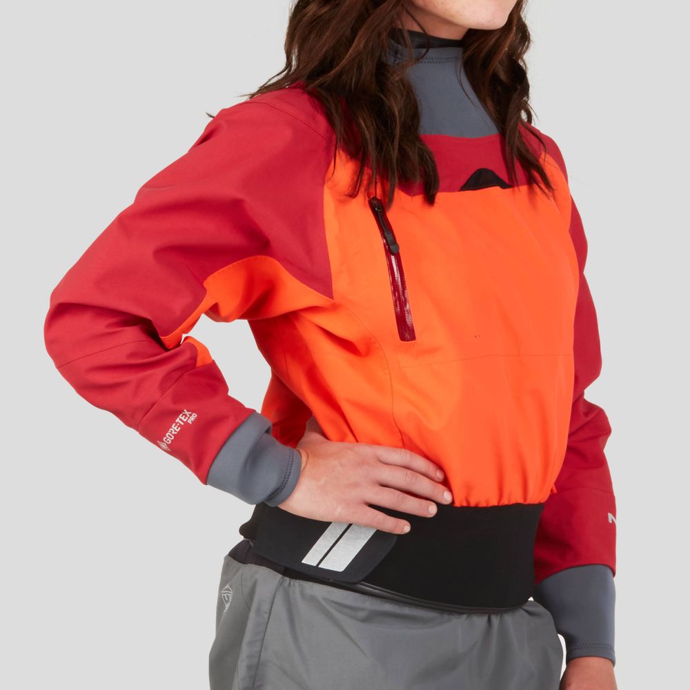 Wearing the REV (GORE-TEX Pro) Drytop by NRS, a red and orange waterproof jacket for paddling with a high collar and black waistband, she confidently stands with one hand on her hip against a plain background.