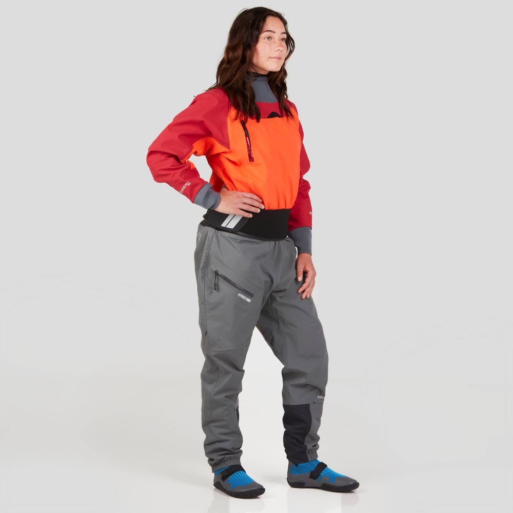 A woman with dark hair stands, wearing the NRS REV (GORE-TEX Pro) Drytop for women in red and orange, paired with gray pants. Her gaze is thoughtful as she enjoys the waterproof breathable performance of her outfit.
