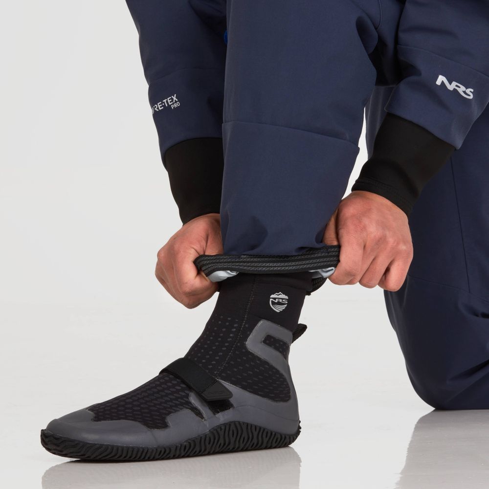 A person adjusts black neoprene water boots while wearing NRS's Axiom (GORE-TEX Pro) Drysuit and dark waterproof pants, all set against a pristine white background.