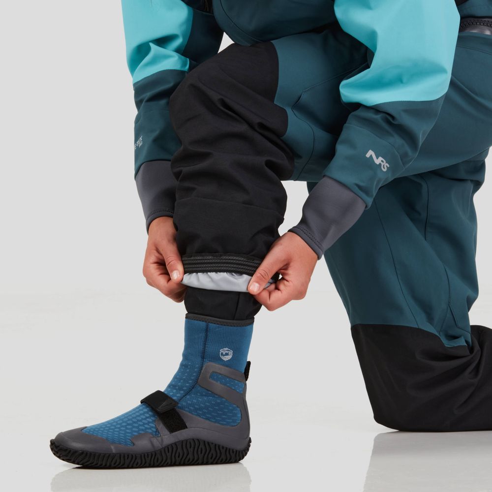 Person kneeling, adjusting the cuff of an NRS Axiom GORE-TEX Pro Drysuit for Women over a blue water shoe, on a white background.