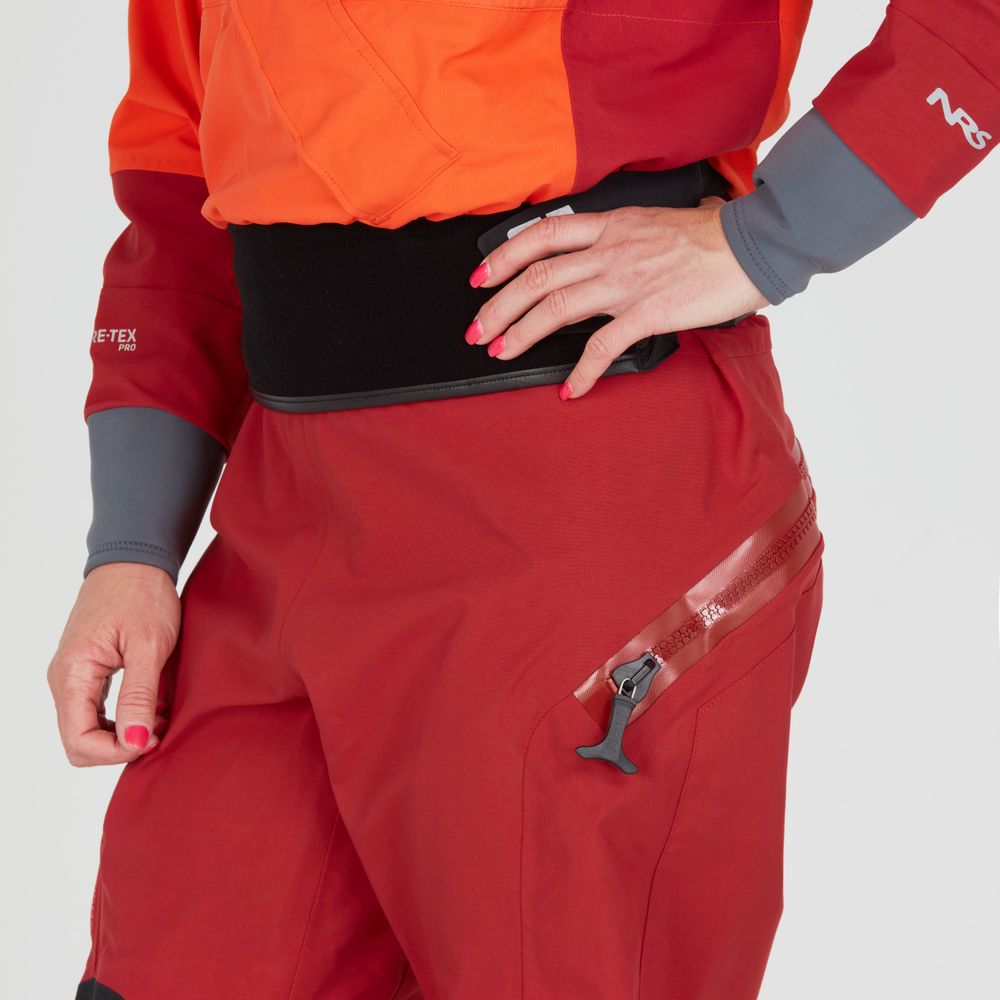 A model poses against a plain backdrop in the NRS Axiom GORE-TEX Pro Drysuit for Women, showcasing its red and orange design with a black waist panel and zippered pocket. This suit offers waterproof, windproof, and breathable protection.
