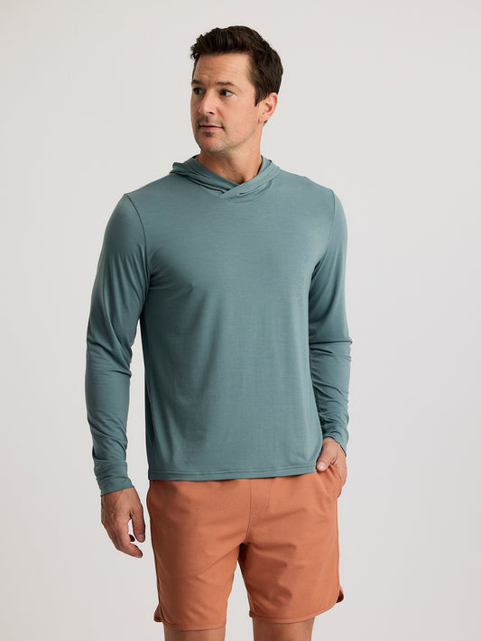 A man in the Free Fly Elevate Lightweight Hoodie M's featuring drirelease technology and light brown shorts stands against a plain light background.