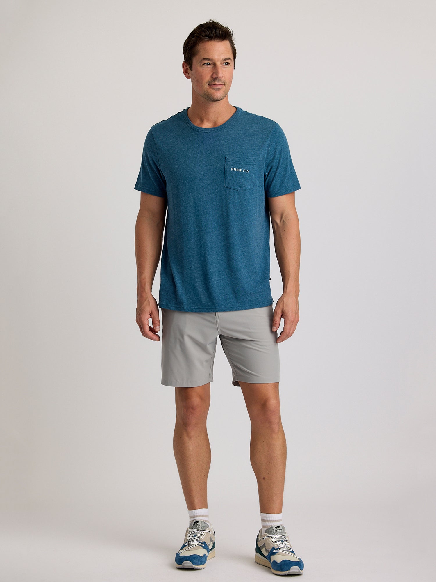 A man faces forward wearing the Free Fly Salt and Sun Pocket Tee M's by Free Fly, paired with light gray shorts and blue and white athletic shoes, set against a plain light background.