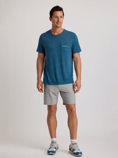 A man faces forward wearing the Free Fly Salt and Sun Pocket Tee M's by Free Fly, paired with light gray shorts and blue and white athletic shoes, set against a plain light background.