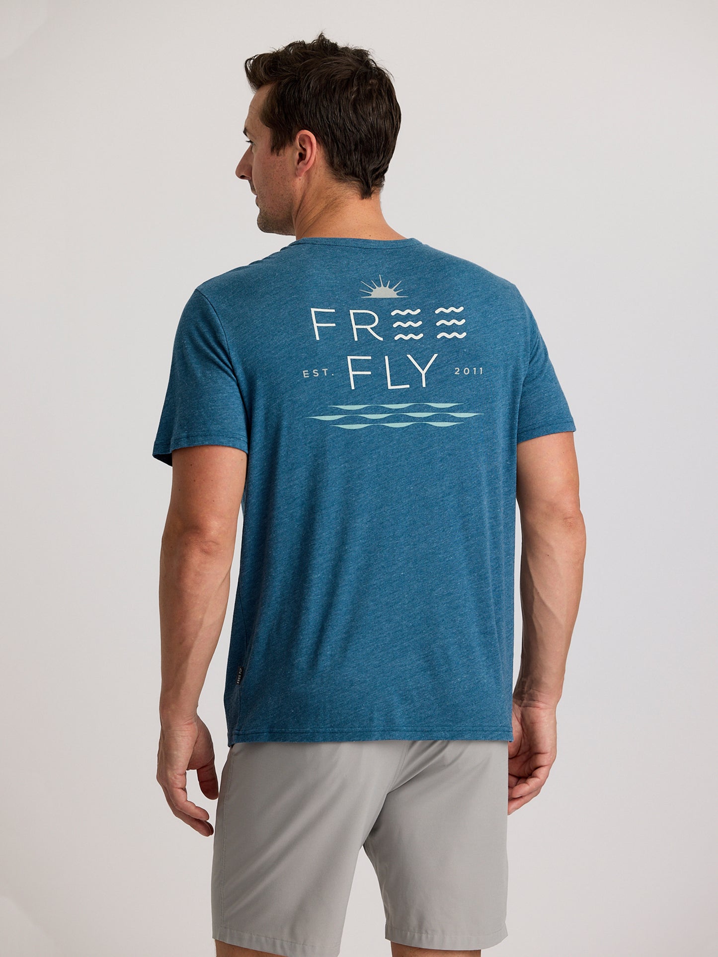 A man faces away wearing the Free Fly Salt and Sun Pocket Tee M's by Free Fly, a blue shirt with “FREE FLY EST. 2011” and wave graphics on the back, paired with light gray shorts.