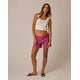 A woman smiles at the camera against a plain background, wearing a white tank top, Carve Oahu 4" Shorts in pink with a matching belt, brown sandals, and a necklace.