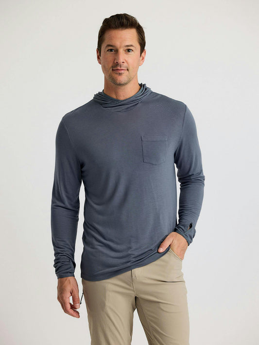 A man stands against a plain background wearing the Free Fly Bamboo Lightweight Hoodie M's by Free Fly, featuring sun protection and a chest pocket, paired with tan pants—ideal for fishing or outdoor adventures.