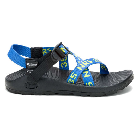 The Chaco Z1 Classic - Men's sandal is designed for outdoor enthusiasts, featuring a textured sole, podiatrist-approved footbed, and adjustable blue and yellow straps with "35 YRS." text and a plastic buckle on the top strap.