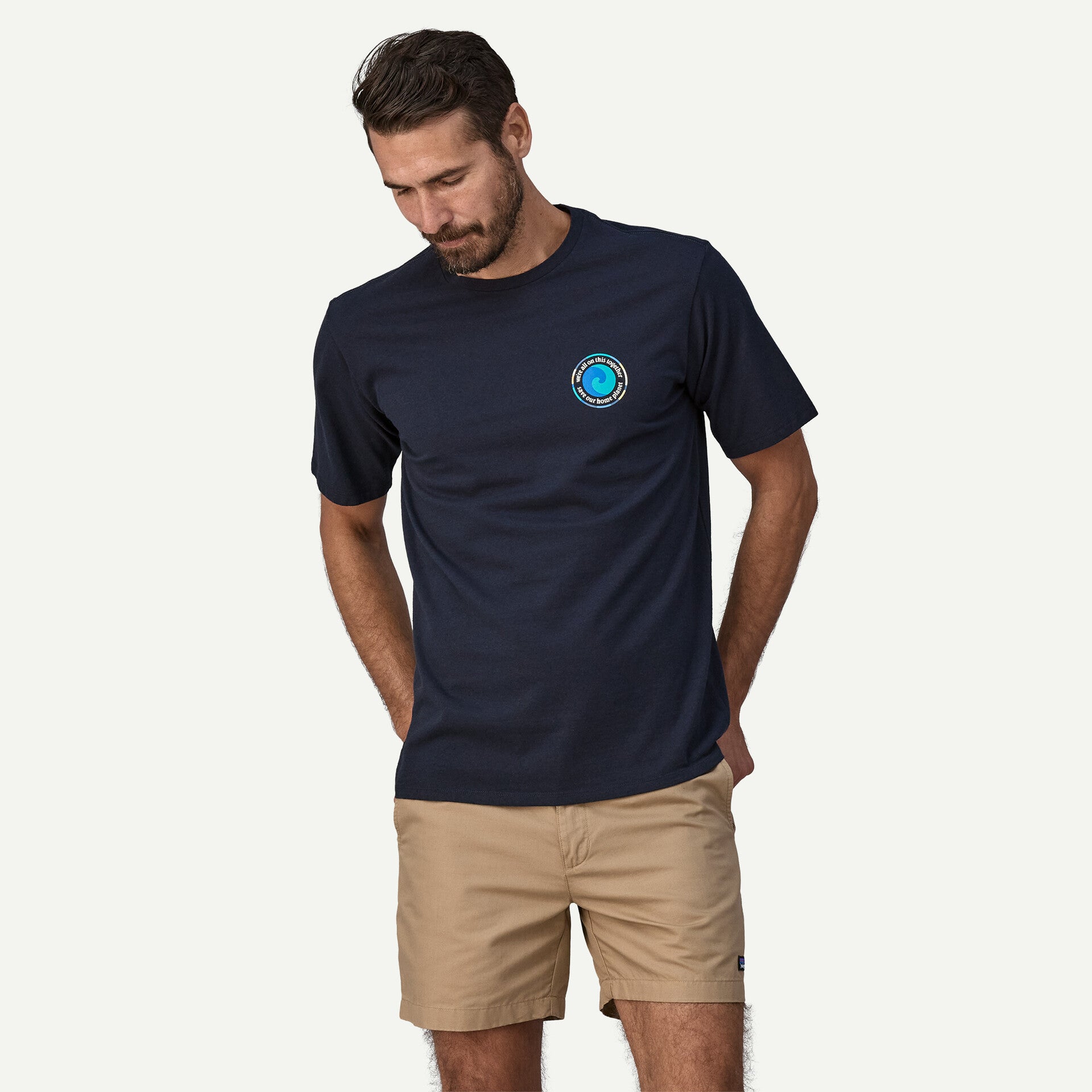 A man wearing a Patagonia Unity Fitz Responsibili-Tee M's in navy and tan shorts stands against a light background, looking down with his hands in his pockets.