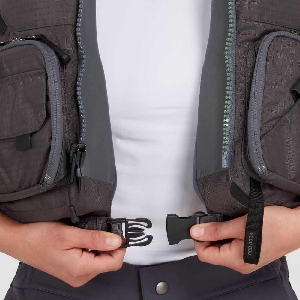 A person fastens the plastic buckle of an NRS Chinook Fishing PFD, featuring two zippered pockets, over a white shirt.