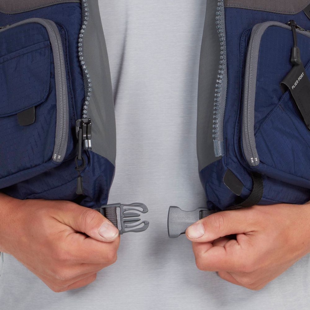 A person fastens the buckle of an NRS Chinook Fishing PFD, a blue and gray life vest with zippered pockets ideal for fishing outings.