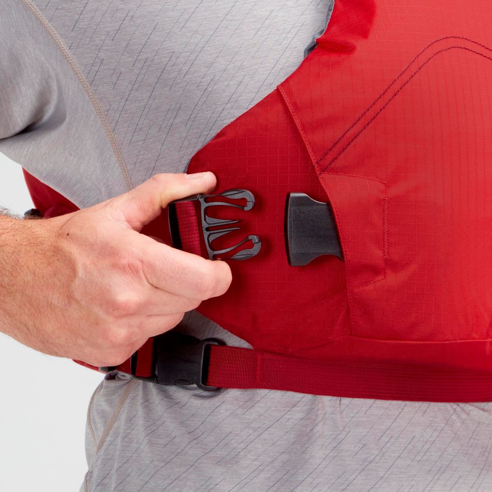 A person in a gray shirt fastens the side buckle of a red NRS Vapor PFD life jacket, preparing to paddle.