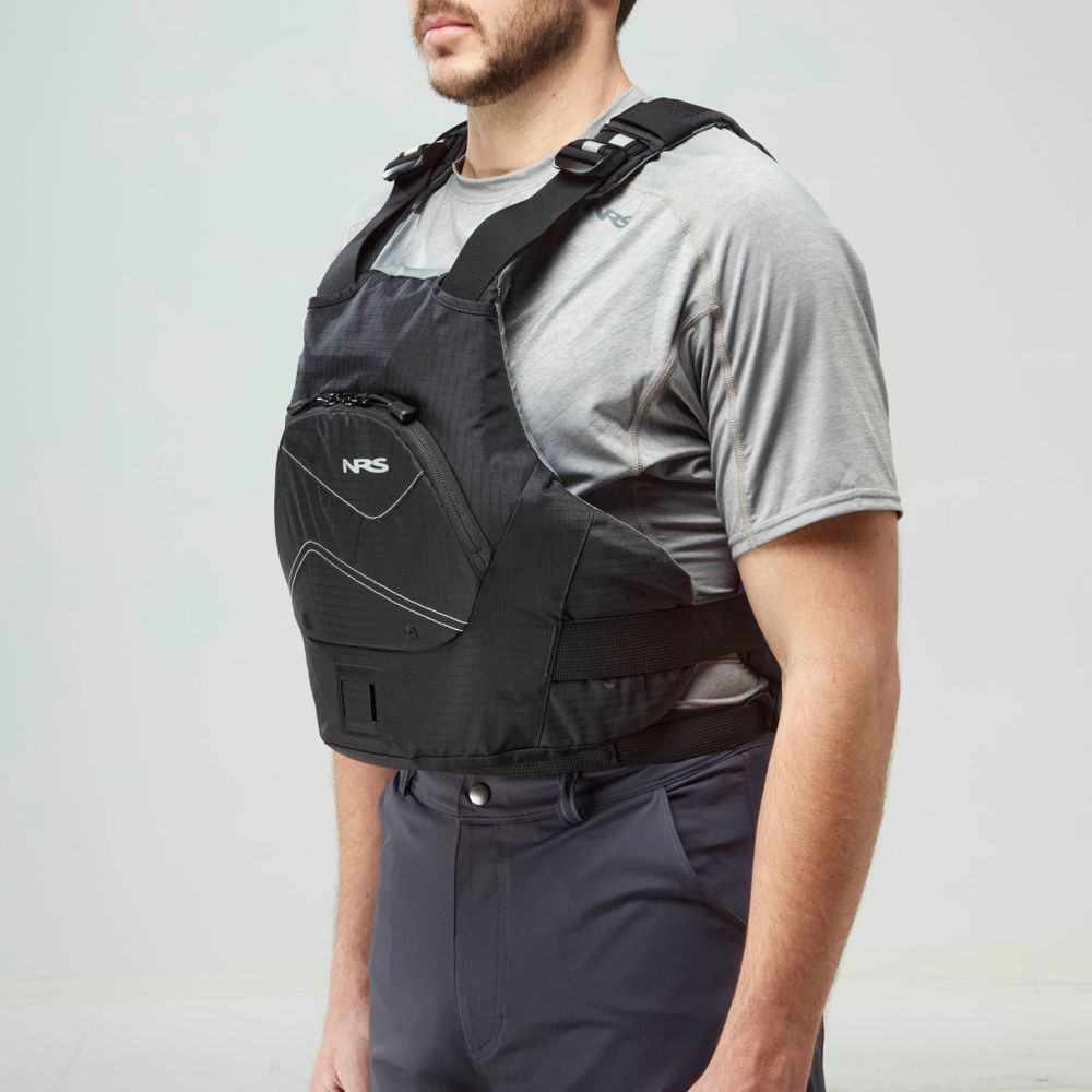 A man models the NRS Ion PFD life jacket in black over a gray short-sleeve shirt and navy pants, set against a plain light background.