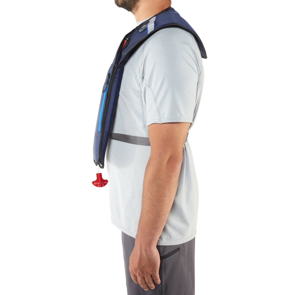 A man in a light gray shirt and dark shorts, wearing the NRS Matik Inflatable PFD, stands in profile view.
