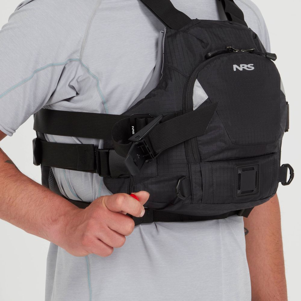 A person in a black NRS Ninja Pro PFD over a light gray shirt holds a safety whistle attached to the vest—perfect for whitewater paddlers.