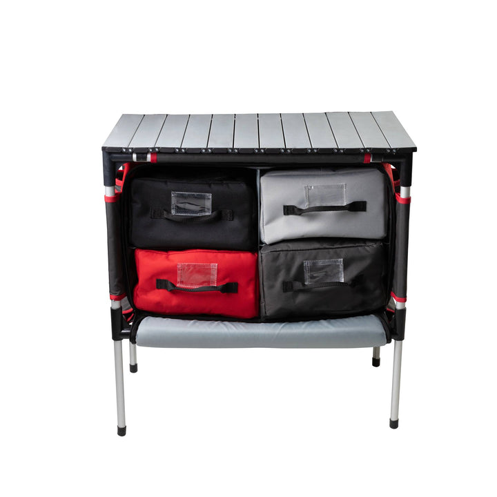 The Camp Chef Sherpa Table & Organizer features a sturdy tabletop and four fabric drawers (two black, one red, one gray) on metal legs—ideal as a camping kitchen or portable organizer for various outdoor setups.