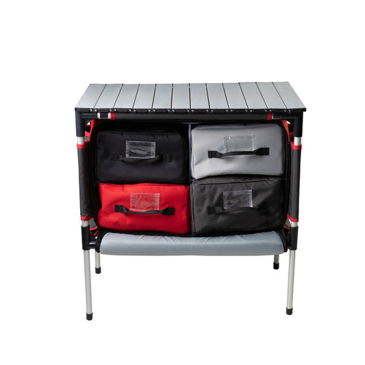 The Camp Chef Sherpa Table & Organizer features a sturdy tabletop and four fabric drawers (two black, one red, one gray) on metal legs—ideal as a camping kitchen or portable organizer for various outdoor setups.