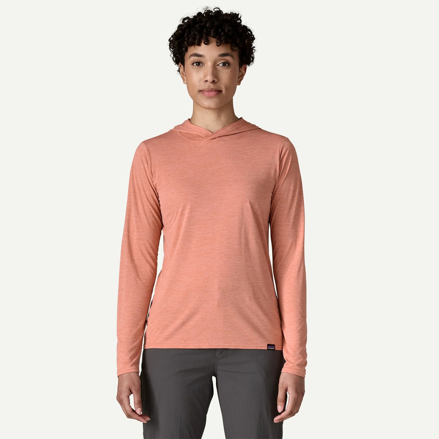 A person wears the Patagonia Cap Cool Daily Hoody for women in light orange, made from recycled polyester, and gray pants, standing facing forward against a plain background.