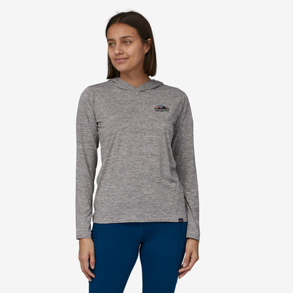 A woman stands against a neutral background, wearing Patagonia's Cap Cool Daily Graphic Hoody, crafted from recycled polyester with a small logo, paired with blue pants.