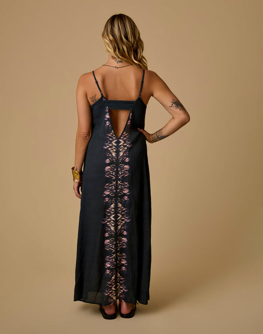 A woman stands with her back to the camera, wearing the Carve Jordy Slip Dress—a black sleeveless maxi with a triangular cutout and tie-dye center pattern.