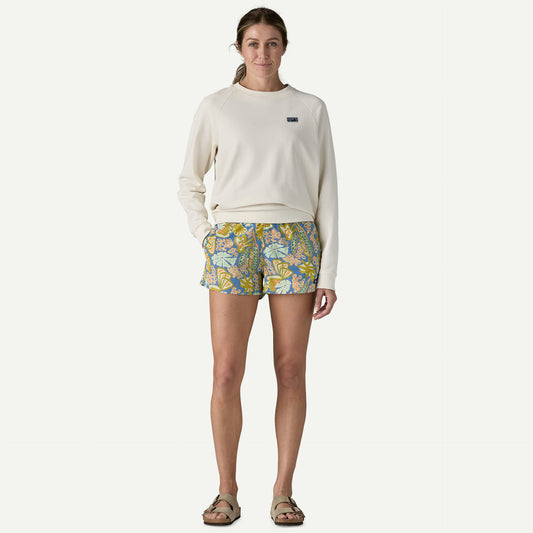 A person wearing a long-sleeve cream sweatshirt, Patagonia Baggies Shorts 2.5 in. - Women's, and sandals stands against a plain background.