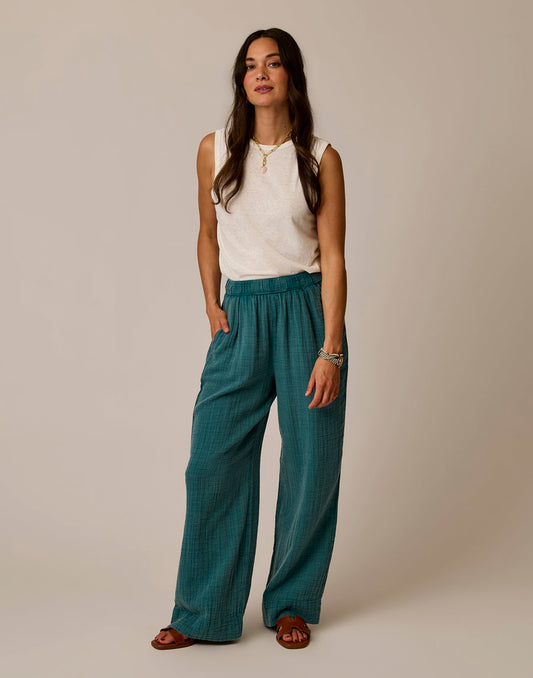 A woman stands against a plain background wearing Carve’s Nico Gauze Pant in teal, a sleeveless white top, and brown sandals, with her hands in her pockets.