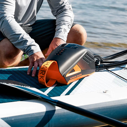 A person is setting up an Outdoor Master Shark 3 - Battery Powered SUP Pump on an inflatable paddleboard near a body of water.