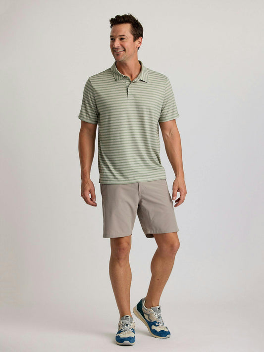 A man wearing a light green striped polo and Free Fly Men's Tradewind Short by Free Fly, paired with blue sneakers, stands smiling against a plain background.