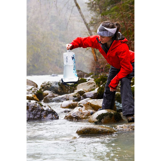 Wearing a red jacket, a person collects water from a rocky stream using the NRS Gravity BeFree Water Filtration System in an outdoor setting.