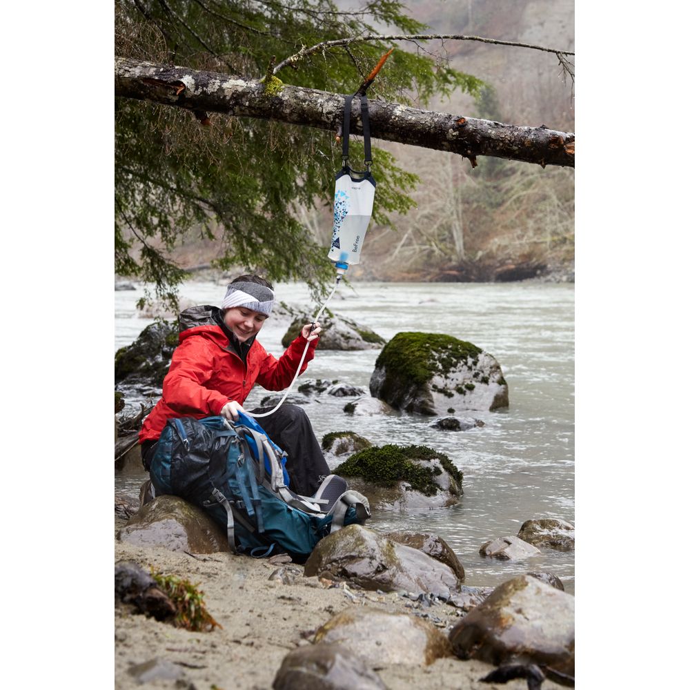 Wearing outdoor gear, a person gathers water from an NRS Gravity BeFree Water Filtration System suspended from a tree branch by a rocky riverbank.