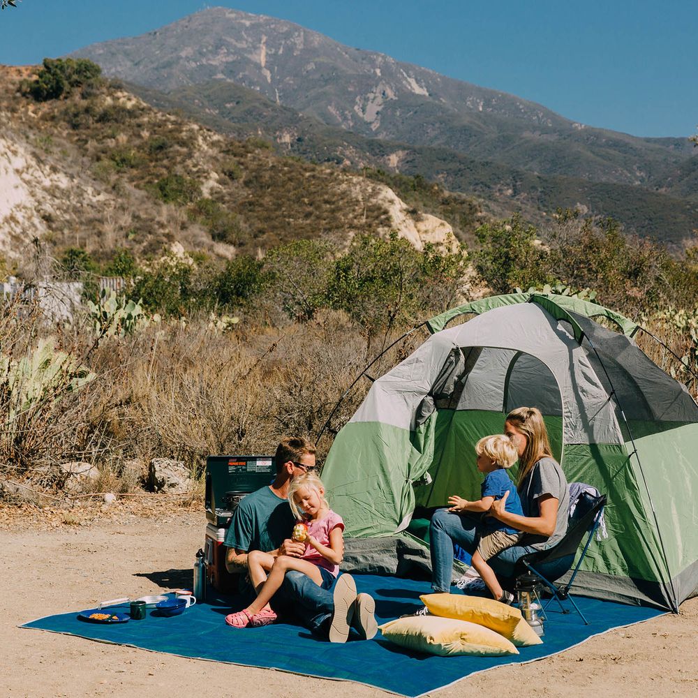 A family of four enjoys a meal on a NRS Sand Free Multi Mat outside their tent, with mountains in the background, relieved of any worry about stray sand interfering with their picnic.