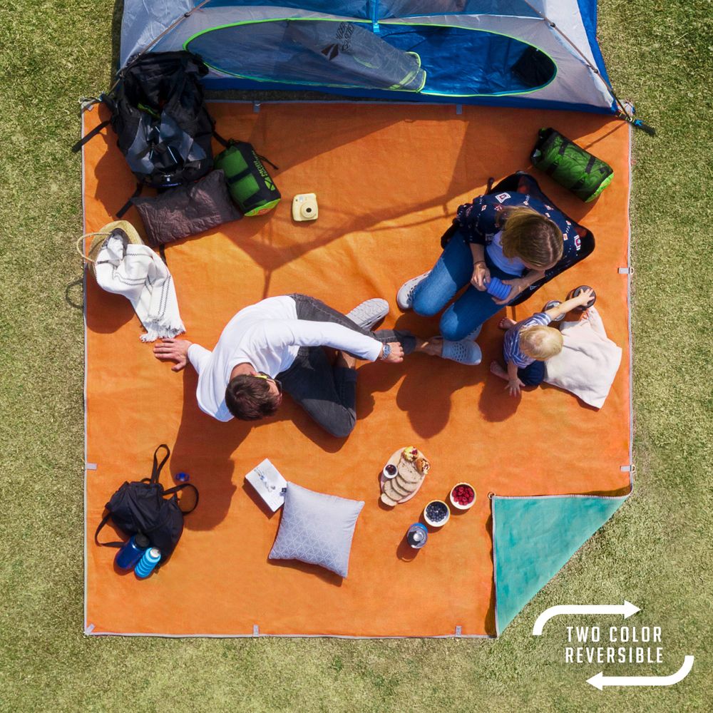 A family enjoys an outdoor adventure on an orange blanket with camping gear, a child, and drinks. The NRS Sand Free Multi Mat's dual weave makes it reversible and keeps the area tidy for a perfect mix of relaxation and practicality.