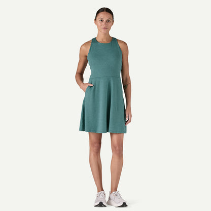 A person wearing a Seabrook Dress by Patagonia—sleeveless and teal with pockets—paired with white sneakers, stands against a plain background.