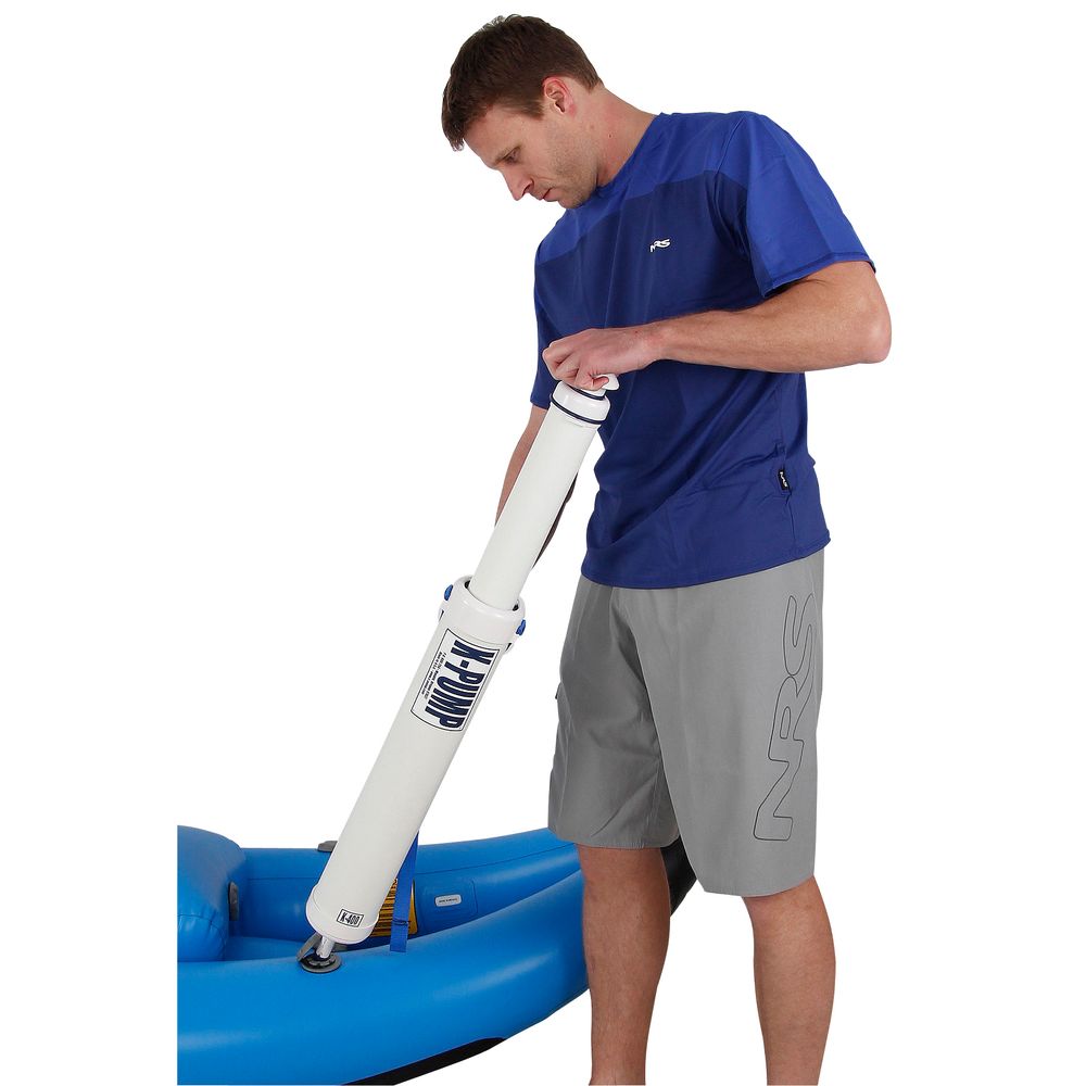 A man in a blue shirt and gray shorts uses the ergonomic NRS K-Pump 400, a white hand pump, to inflate an inflatable blue kayak.