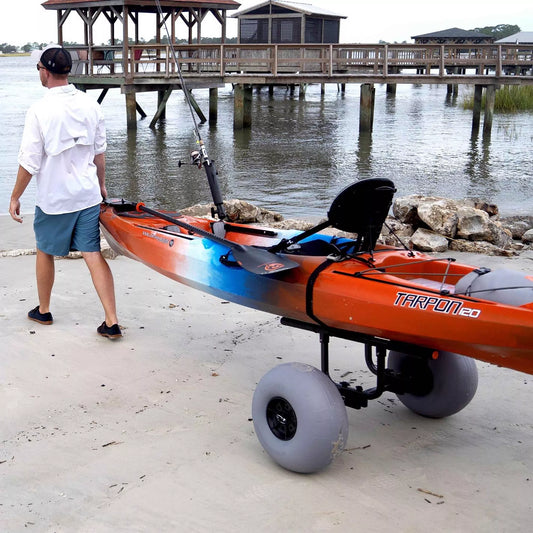 A person pulls their kayak on a Wilderness Systems Heavy Duty Kayak Cart with 13" beach wheels, effortlessly cruising along the sandy shore as the dock and sparkling water shimmer in the background.