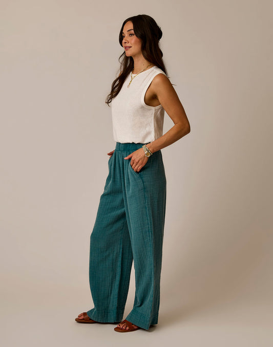 A woman stands in profile against a plain background, wearing Carve's Nico Gauze Pant in teal with a sleeveless white top, sandals, and jewelry, her hands tucked into her pockets.