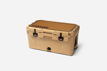 A tan hard plastic cooler with black latches and handles features a Canyon Coolers Cooler Traction Pad made of EVA foam, labeled "Canyon" on the lid and front, shown against a plain white background.