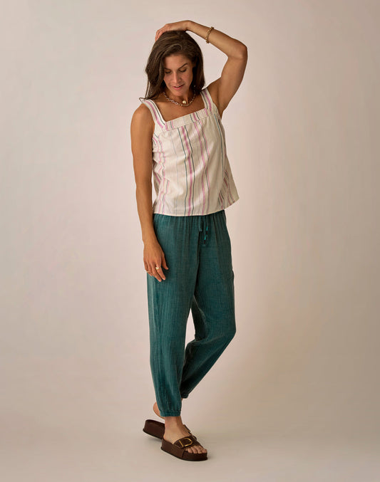 A woman stands against a plain background wearing Carve’s Calista Gauze Pant with a striped sleeveless top, brown sandals, minimal jewelry, and one hand touching her hair.