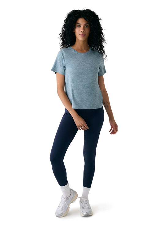 A person with curly hair stands facing forward, wearing the light blue Vital Tee from Lole and navy sustainable activewear leggings, paired with white socks and sneakers against a plain white background.