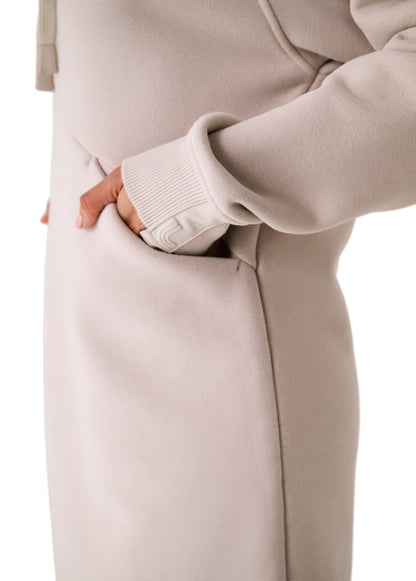A person wears the Lole Yonder Hoodie Dress in beige, with their right hand inside the side pocket; only the torso and arm are visible.