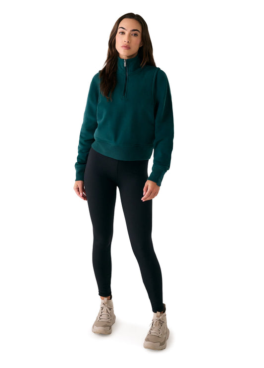 A woman stands against a white background wearing the Lole Yonder Half Zip in teal, black leggings, and beige sneakers.