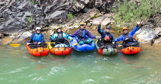 Five people in helmets and life jackets pose in four inflatable kayaks on a river near grassy banks in Durango, CO during their ACA Packraft Instructor Certification with 4CRS Paddle School.