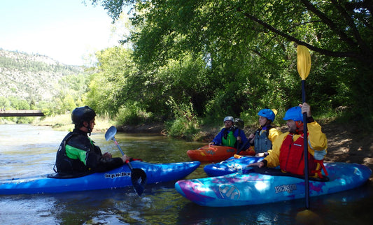 Four adults in helmets and life jackets learn kayaking basics on a sunny riverside as part of the 4CRS Paddle School’s Adult Level 1 Kayak Class.