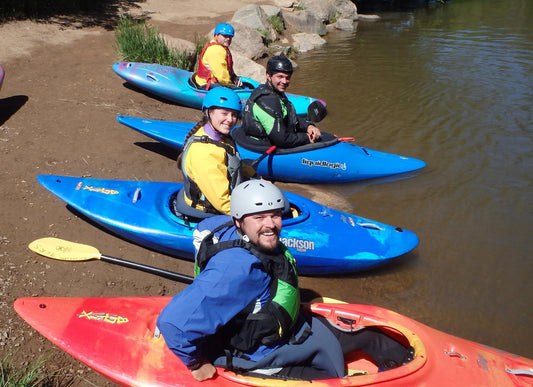 Four adults in kayaking gear smile from colorful kayaks onshore, ready for adventure—an ideal moment to gift the 4CRS Adult Paddle School Class Holiday Gift Certificate for a fun day of paddling.